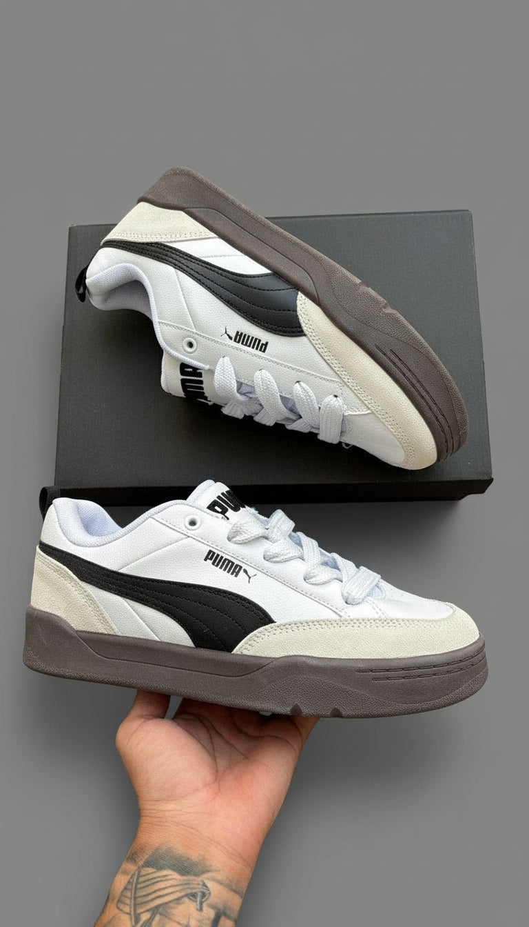 White and black sneakers with brown soles held by a hand on a wooden surface.